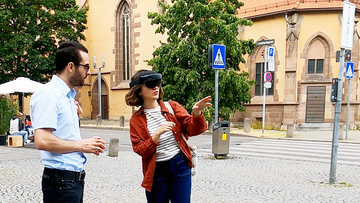 Eine Frau steht mit einer VR-Brille in der Leonhardsvorstadt und lässt sich von einem Mann die Anwendung erklären, im Hintergrund die Leonhardskirche